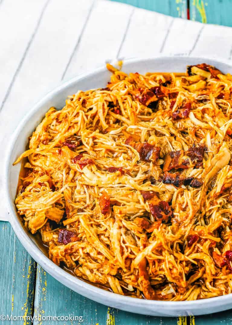 A white plate filled with super easy Bacon Barbecue Chicken—shredded barbecue chicken topped with crispy bacon—rests on a striped cloth over a wooden table.