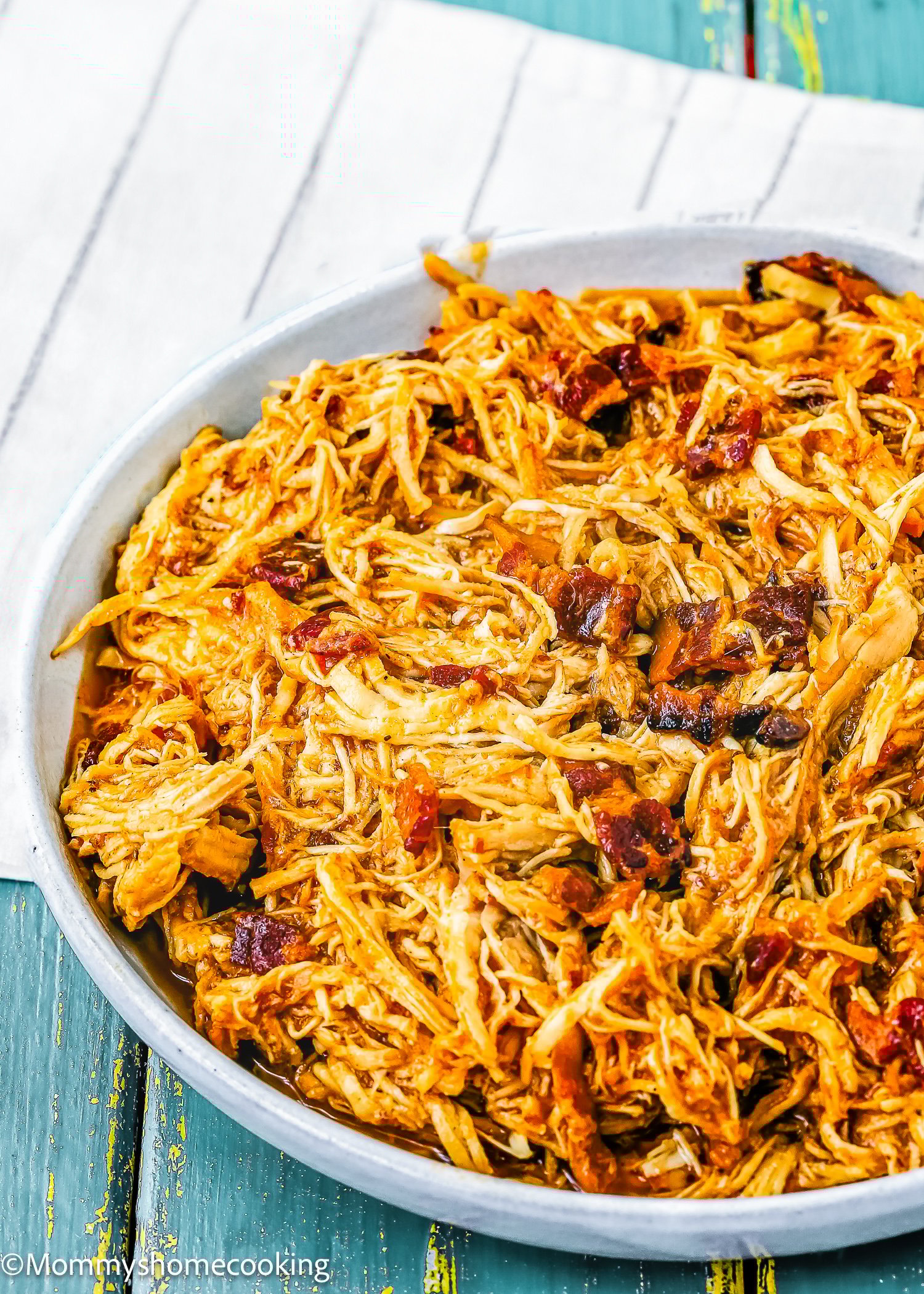 A white plate filled with super easy Bacon Barbecue Chicken—shredded barbecue chicken topped with crispy bacon—rests on a striped cloth over a wooden table.