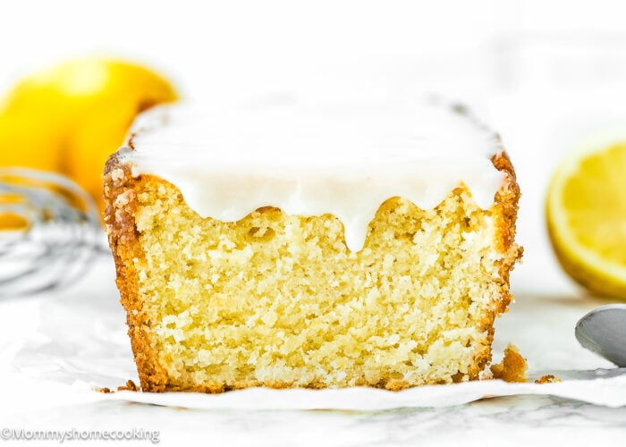 A close-up of a slice of lemon loaf cake with white icing on top, showing a moist, dense texture—just like you’d expect from an Easy Eggless Lemon Pound Cake Recipe (No Eggs!). A halved lemon and a whisk are visible in the background.