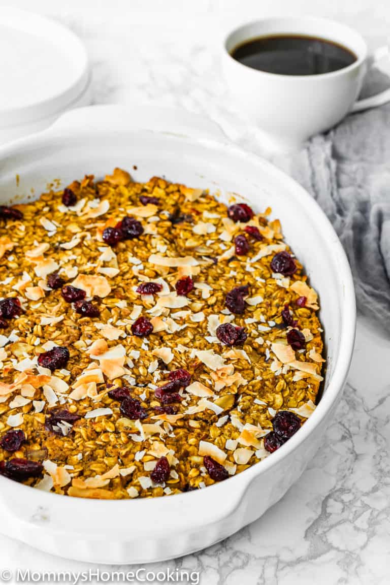A white baking dish filled with Eggless Baked Pumpkin Oatmeal, topped with dried cranberries and coconut flakes, sits on a marble surface next to a cup of black coffee.