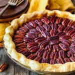 egg-free pecan pie in a pie dish over a wooden surface with a yellow kitchen towel, wooden plates and forks in the background.