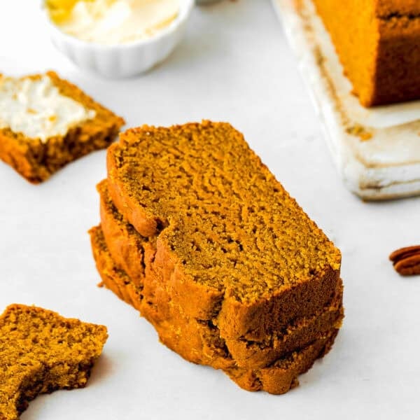 Three slices of eggless pumpkin bread stacked on a white surface, with a buttery slice and a bitten piece nearby. A butter dish and pecans complete the cozy scene in the background.