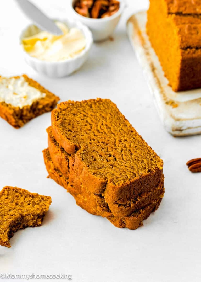 Three slices of eggless pumpkin bread stacked on a white surface, with a buttery slice and a bitten piece nearby. A butter dish and pecans complete the cozy scene in the background.