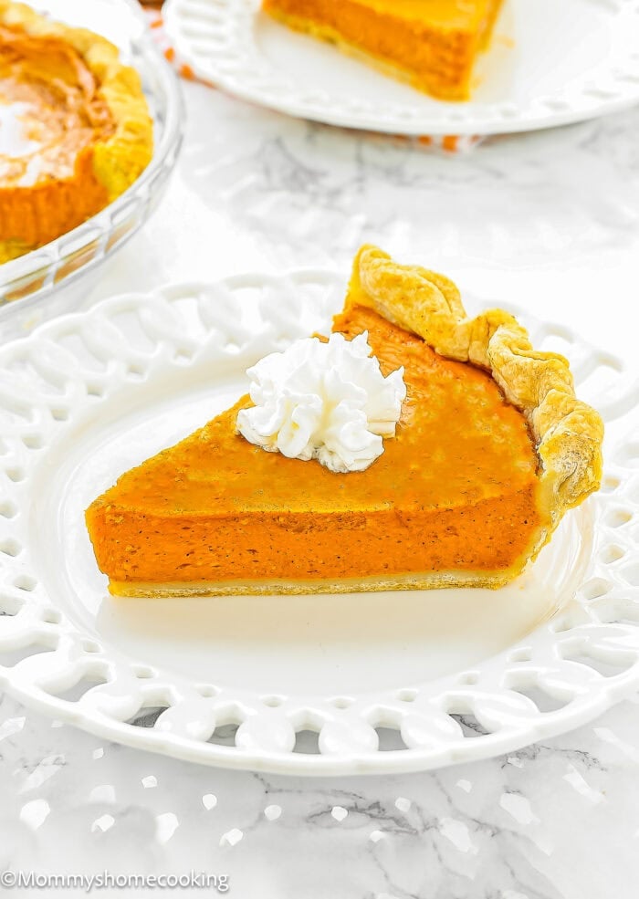 A slice of Pumpkin Pie without eggs with a dollop of whipped cream on top, served on a white decorative plate.