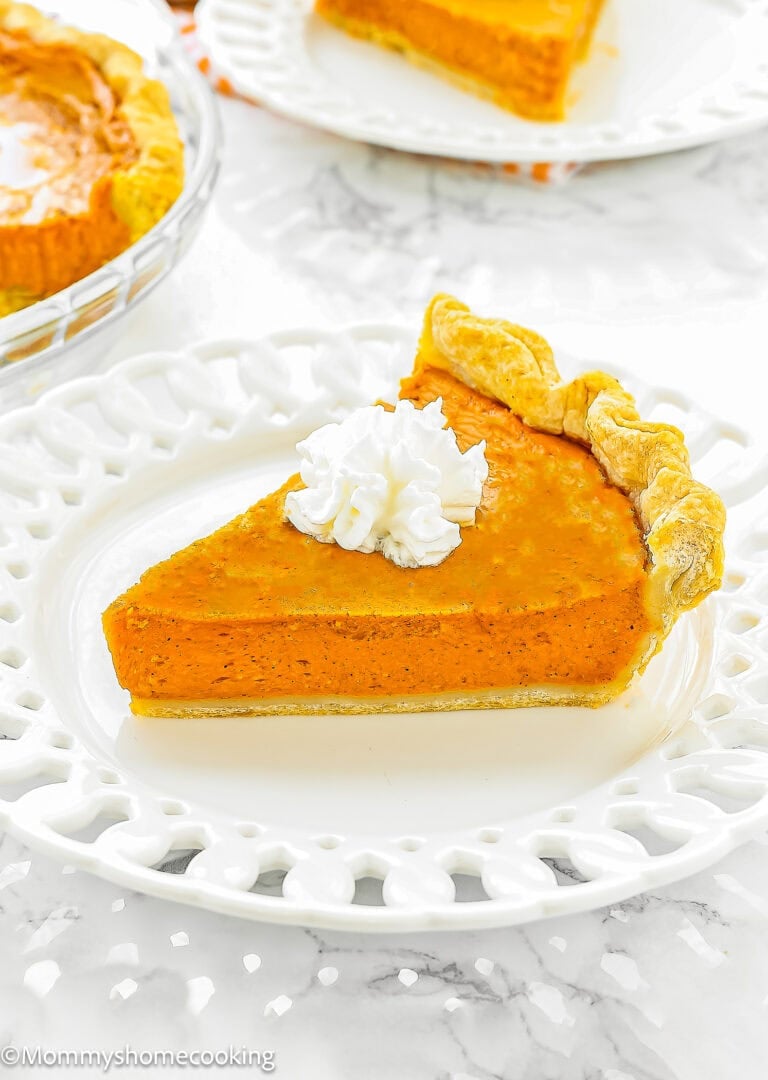 A slice of Pumpkin Pie without eggs with a dollop of whipped cream on top, served on a white decorative plate.
