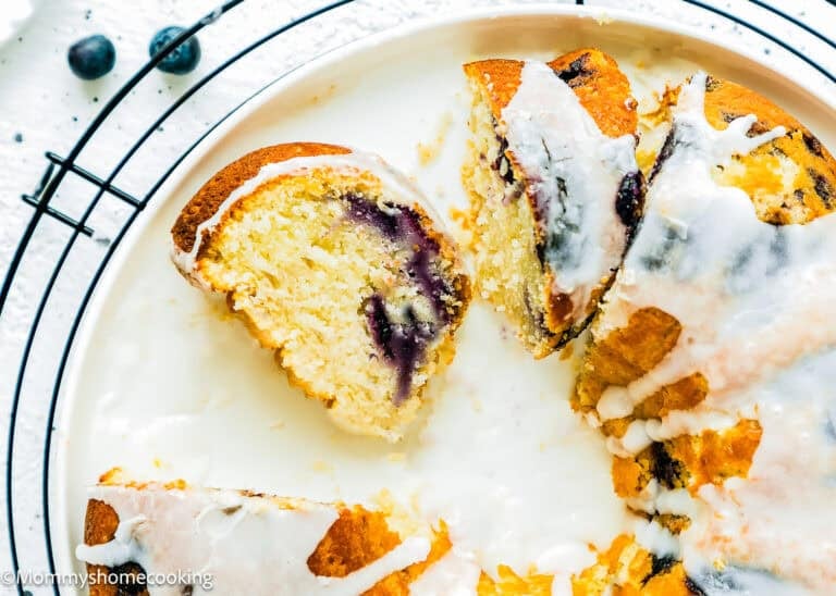 A partially sliced Eggless Lemon Blueberry Bundt Cake with white icing on top, placed on a white plate with a cooling rack underneath.
