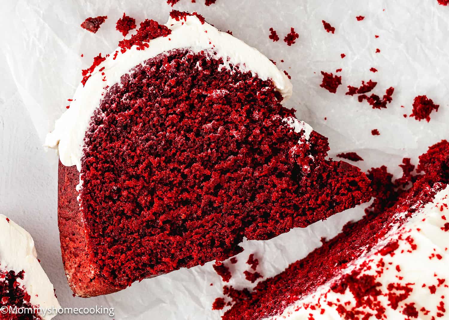 A slice of Eggless Red Velvet Bundt Cake with cream cheese frosting sits on white parchment paper, surrounded by scattered cake crumbs.