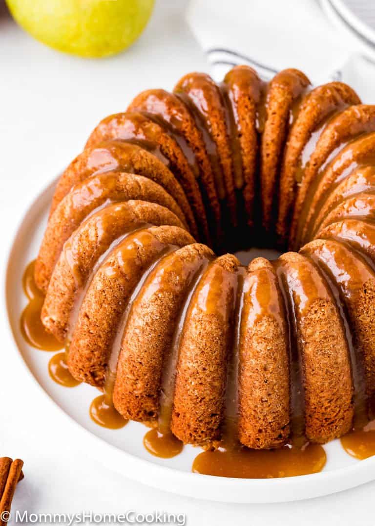An Eggless Apple Cider Donut Cake drizzled with caramel sauce sits on a white plate, with an apple and cinnamon sticks visible in the background.