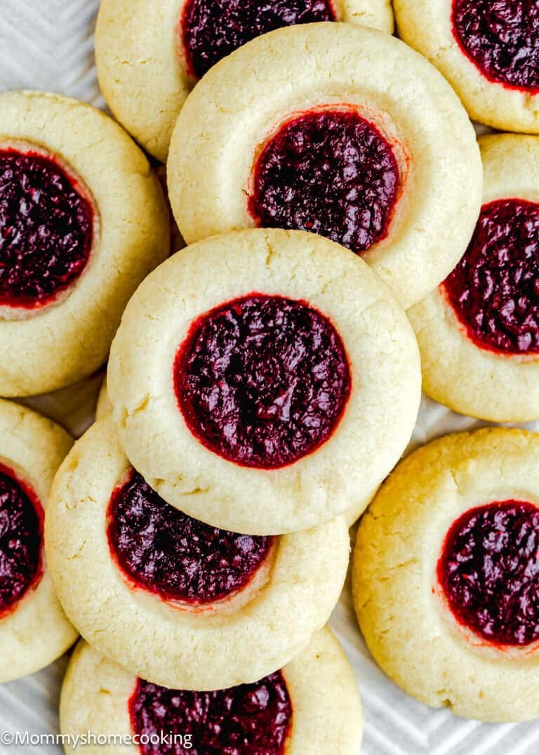 Close-up of several Eggless Thumbprint Cookies with a light-colored dough and a red jam filling in the center, arranged in an overlapping pile.