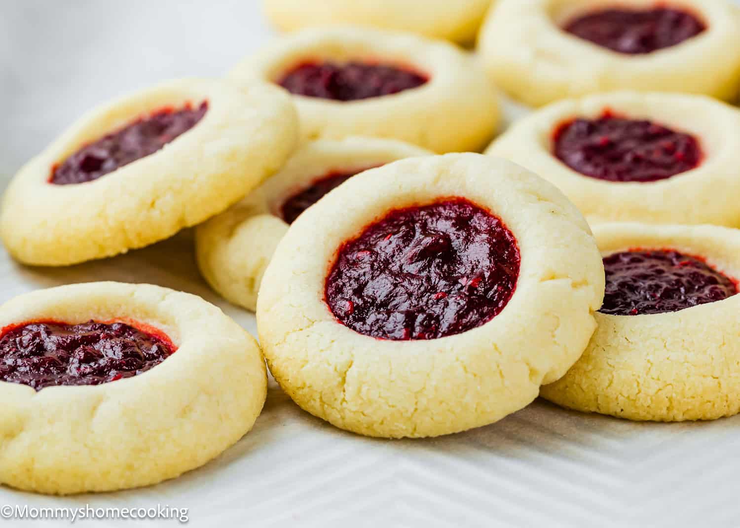 A close-up of several Eggless Thumbprint Cookies with a red jam filling, arranged on a white surface.