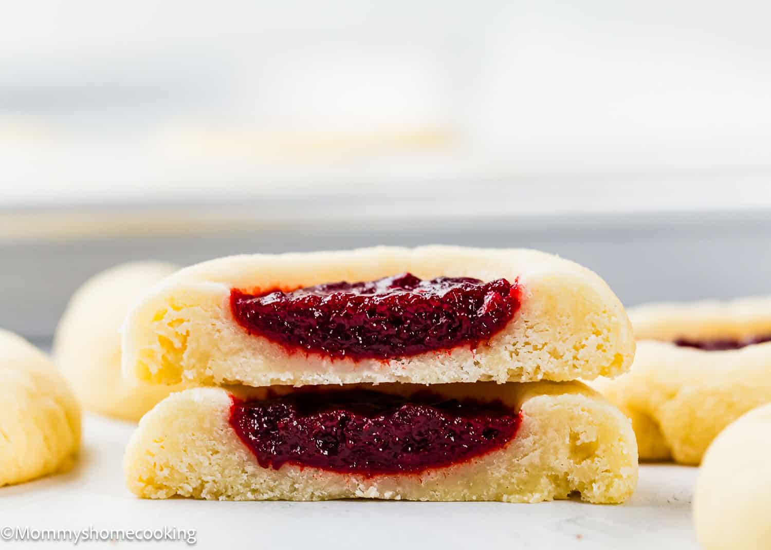 Close-up of two Eggless Thumbprint Cookies filled with red jam, one stacked on top of the other and cut in half to reveal the luscious filling.