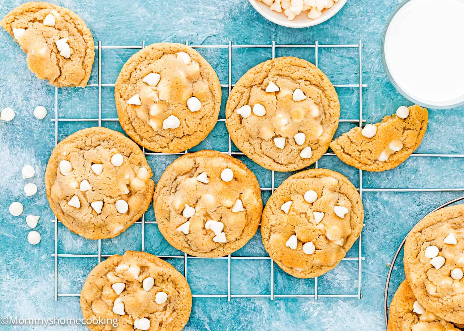 Eggless white chocolate cookies with creamy chips, cooling on a wire rack next to a glass of milk on a blue surface.