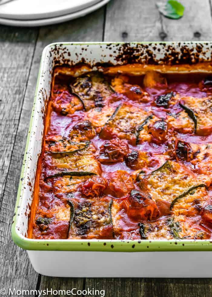 Baked eggless lasagna zucchini rolls in a rich tomato sauce, sprinkled with cheese, nestled in a rectangular ceramic dish on a wooden surface.