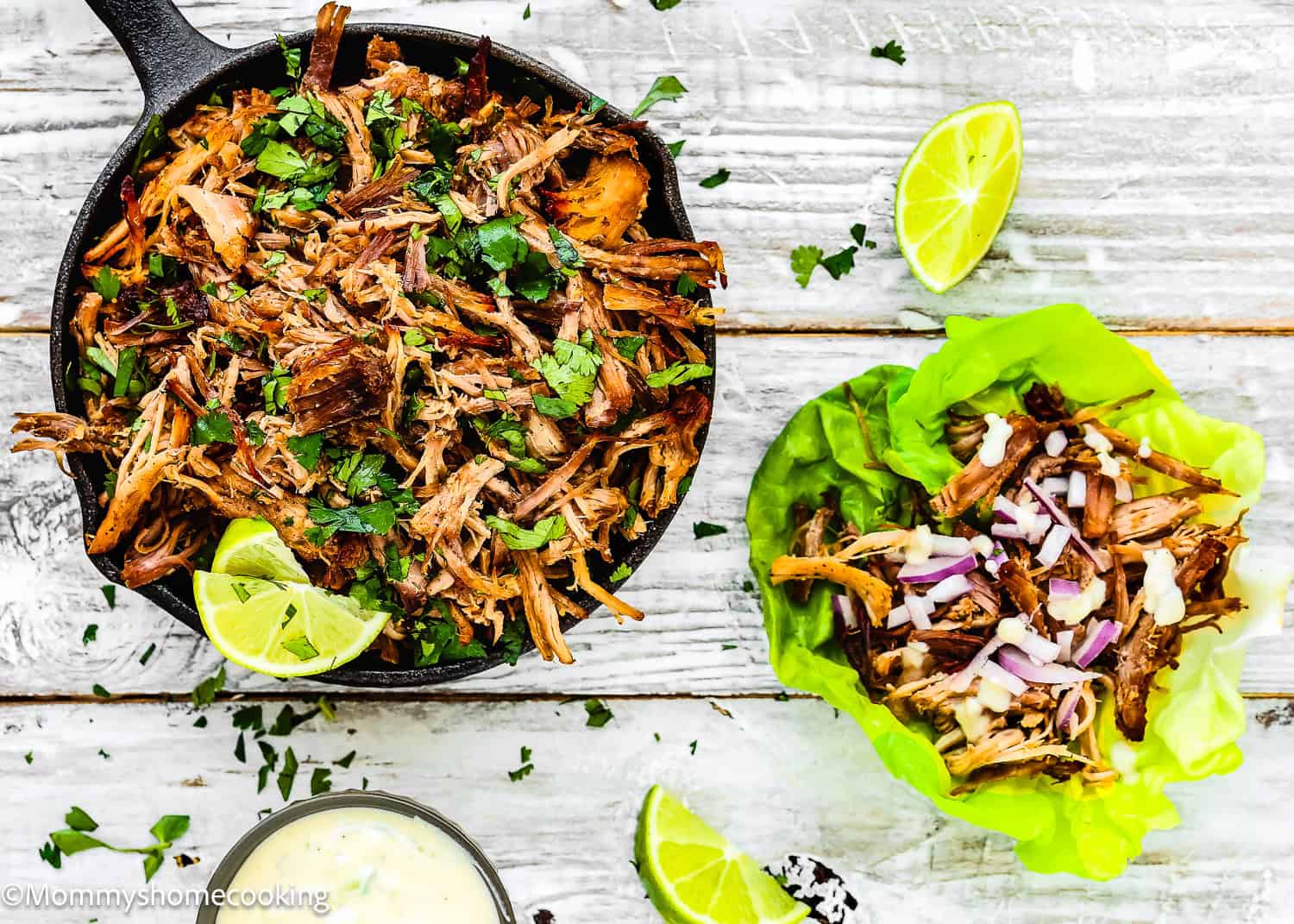 Instant Pot Crispy Pork Carnitas are shredded to perfection, garnished with cilantro in a pan and nestled in lettuce wraps. Lime wedges on the side add zest, all presented beautifully on a wooden surface.