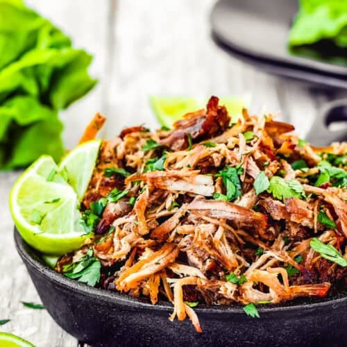 A plate of shredded and cooked Instant Pot crispy pork carnitas is beautifully adorned with chopped herbs, lime wedges, and fresh lettuce in the background on a rustic wooden surface.