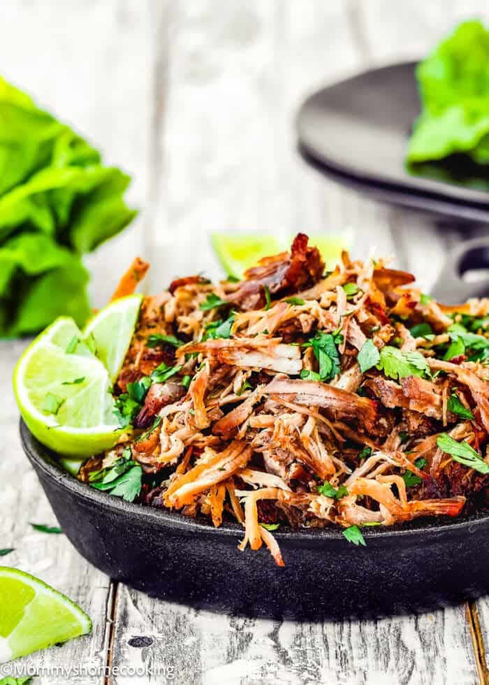 A plate of shredded and cooked Instant Pot crispy pork carnitas is beautifully adorned with chopped herbs, lime wedges, and fresh lettuce in the background on a rustic wooden surface.
