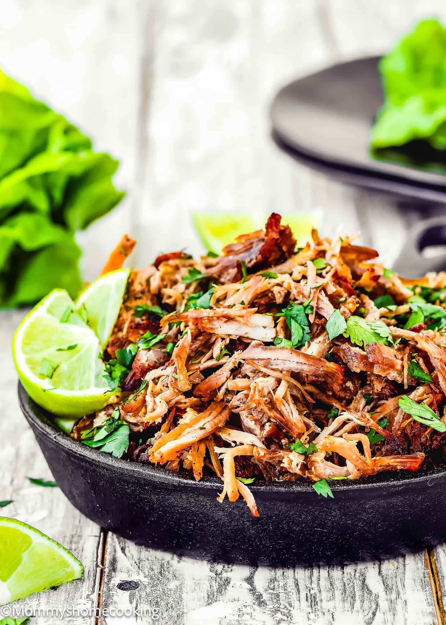 A cast iron skillet brimming with Instant Pot Crispy Pork Carnitas is garnished with parsley and lime wedges, resting on a wooden table. In the background, lettuce leaves blur alongside another skillet, adding to the rustic charm of the scene.