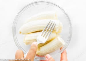 A hand holding a fork is about to mash peeled bananas in a clear glass bowl on a white surface, preparing the base for the BEST Eggless Banana Bread.