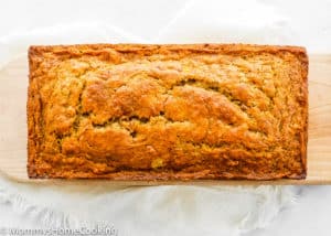 A loaf of golden-brown BEST Eggless Banana Bread sits on a wooden board, resting on a white cloth.