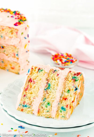 eggless funfetti cake slice on a plate with the whole cake in the background.