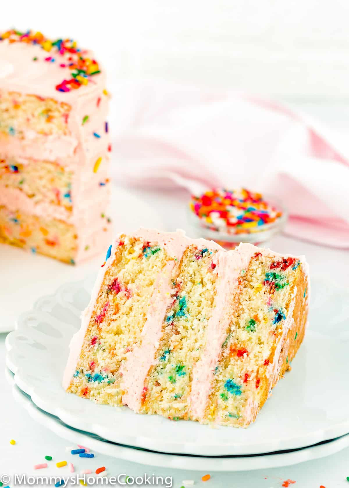 eggless funfetti cake slice on a plate with the whole cake in the background.