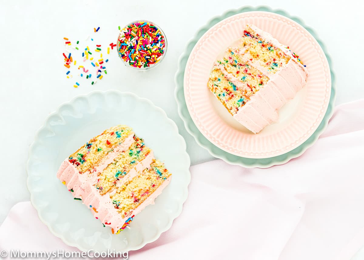 two slices of eggless funfetti cake on plates with a pink kitchen towel and sprinkles on the side.