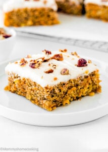 A slice of Eggless Pineapple Carrot Sheet Cake topped with white frosting and chopped nuts on a white plate.