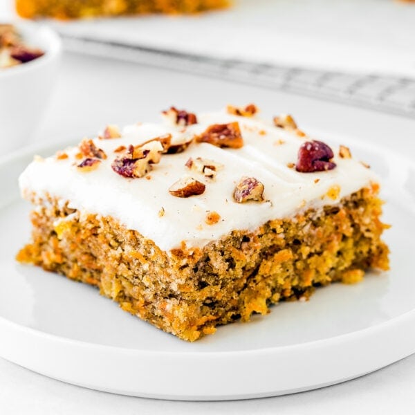 A slice of Eggless Pineapple Carrot Sheet Cake topped with white frosting and chopped nuts on a white plate.