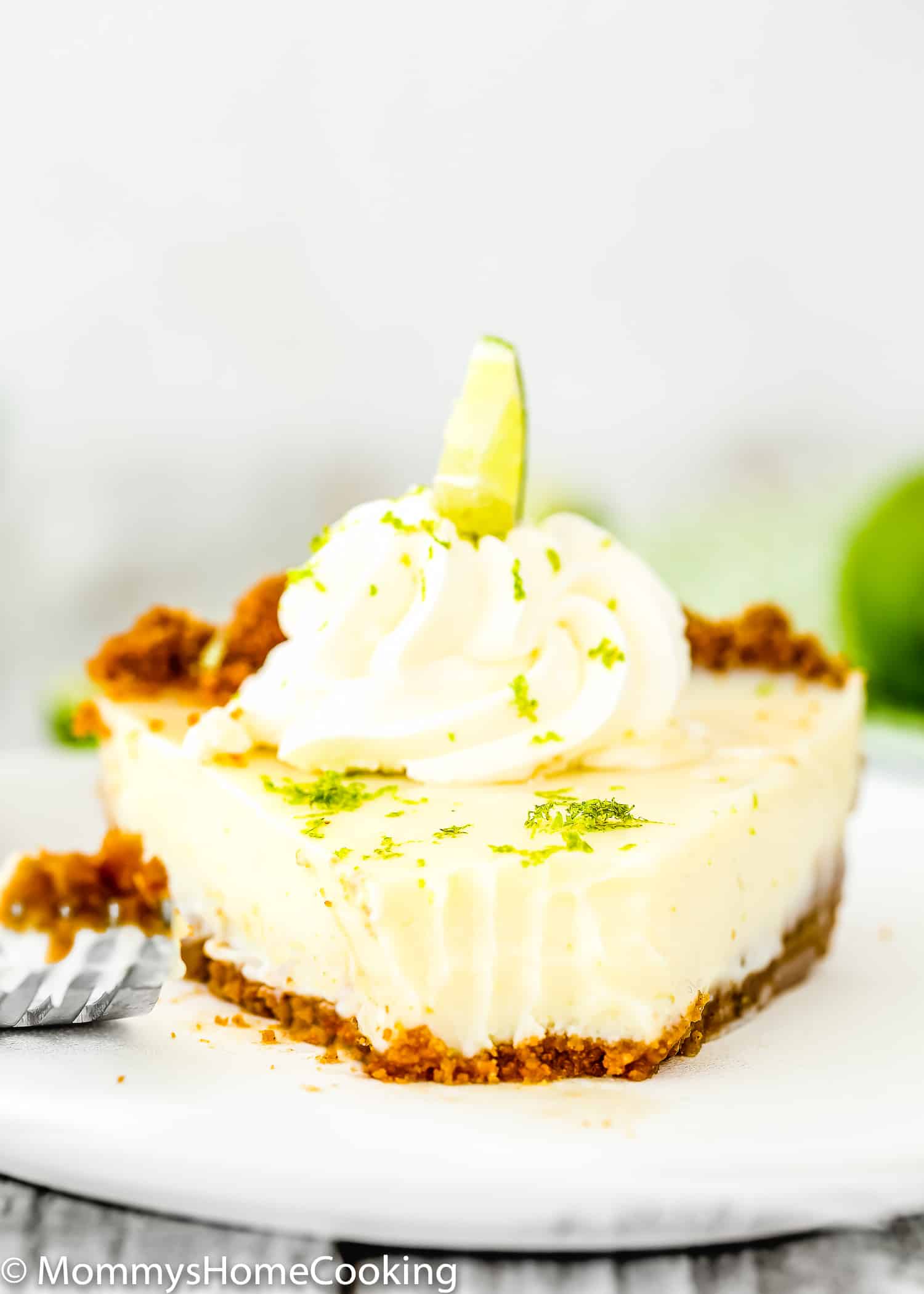 slice of egg-free key lime pie with whipped cream and fresh lime zest on top in a plate with a fork on the side.