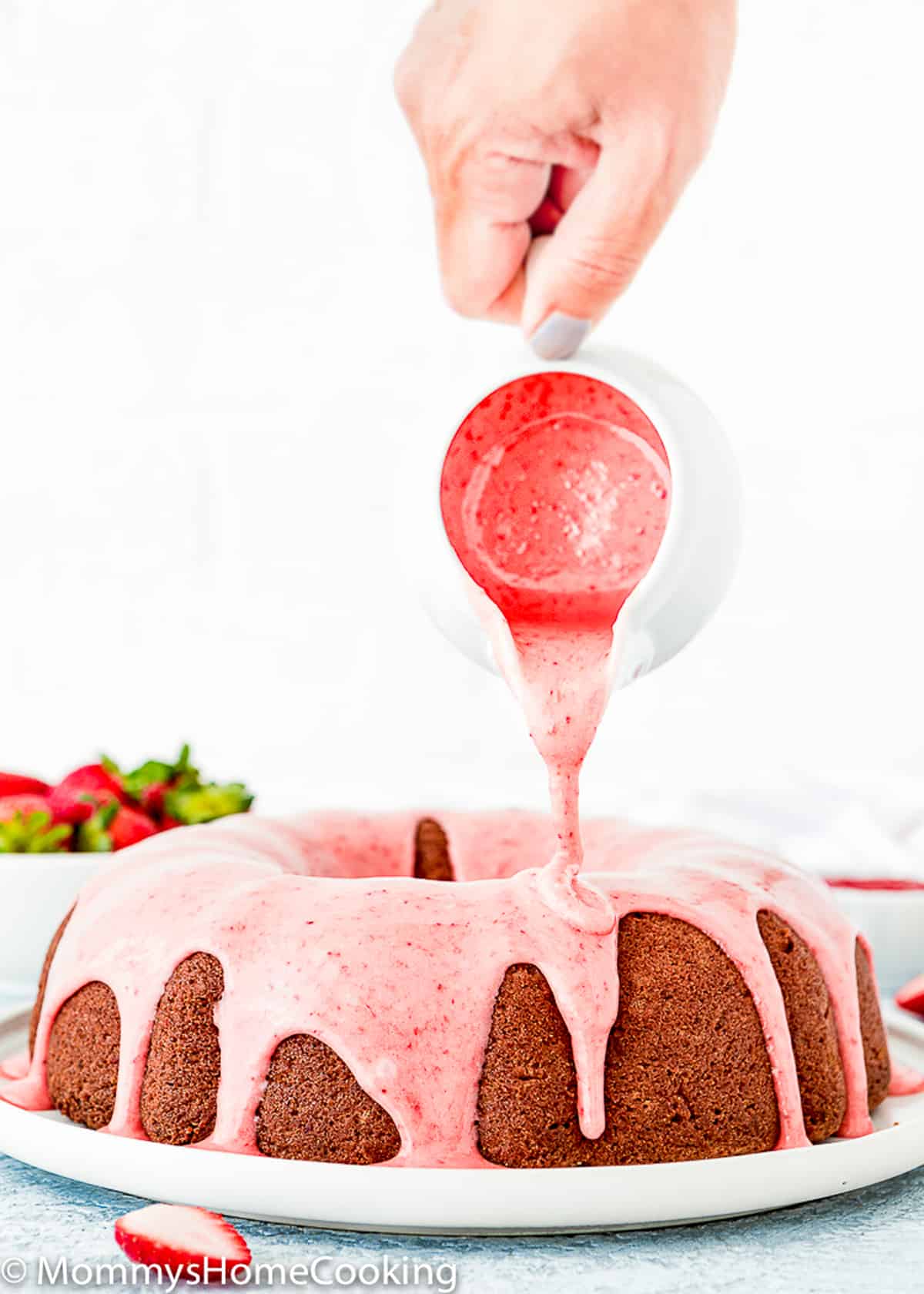 a hand pouring strawberry glaze over an Eggless Homemade Strawberry Bundt Cake on serving plate.