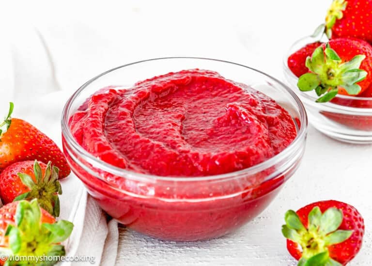 A glass bowl filled with smooth strawberry puree sits on a white surface, surrounded by fresh whole strawberries, highlighting the vibrant color and luscious texture of the strawberry puree.