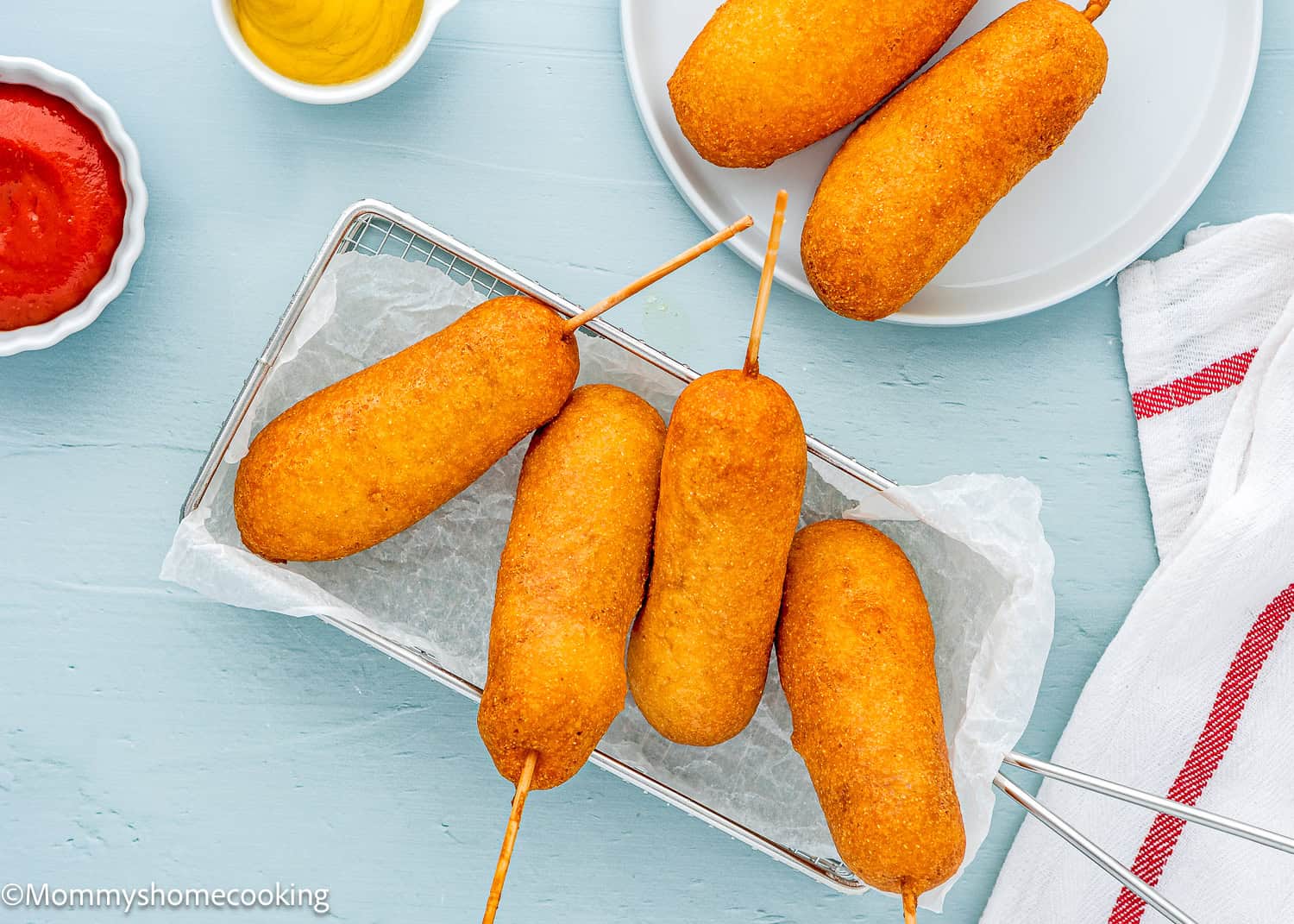 Five of The Best Eggless Corn Dogs (Fair Food at Home) on sticks are arranged on a tray, with two small bowls of ketchup and mustard alongside, while a plate holding two more corn dogs sits nearby, all set on a light blue surface with a white and red towel.