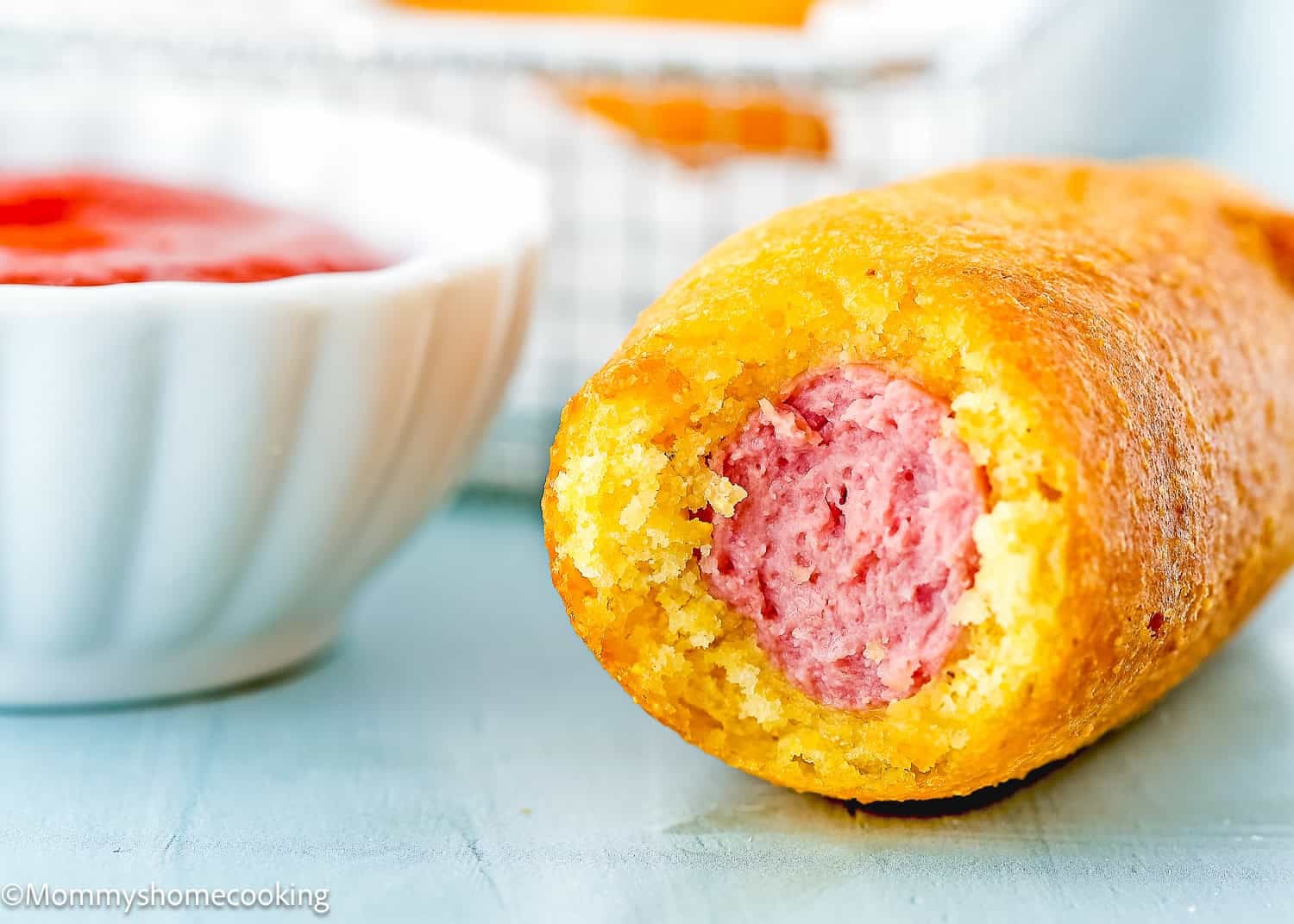 A close-up of a bitten corn dog from The Best Eggless Corn Dogs (Fair Food at Home) shows the sausage inside, with a bowl of red dipping sauce in the background.