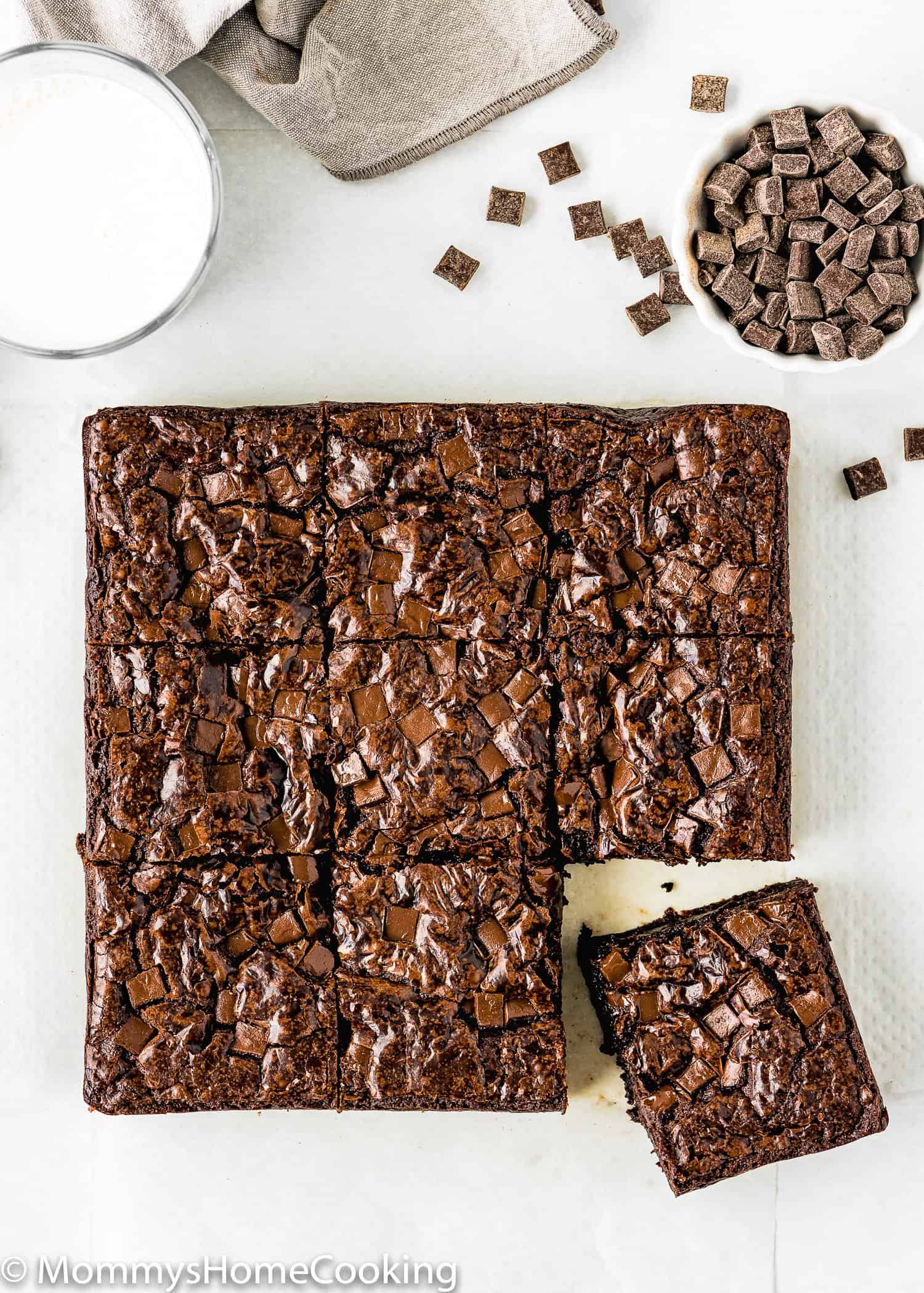 egg-free brownies cut into squares over a white surface with a glass of milk and chocolate chunks on the side.