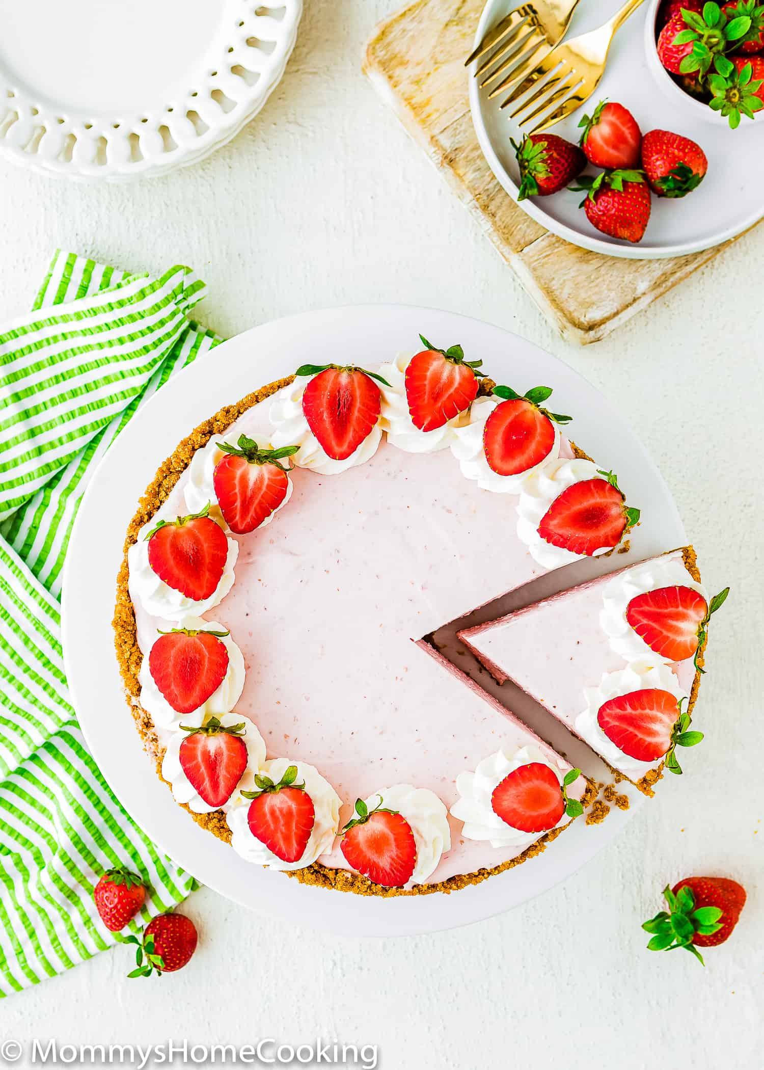cut No-Bake Strawberry Cheesecake over a cake stand with white plates on the side and a green kitchen towel.