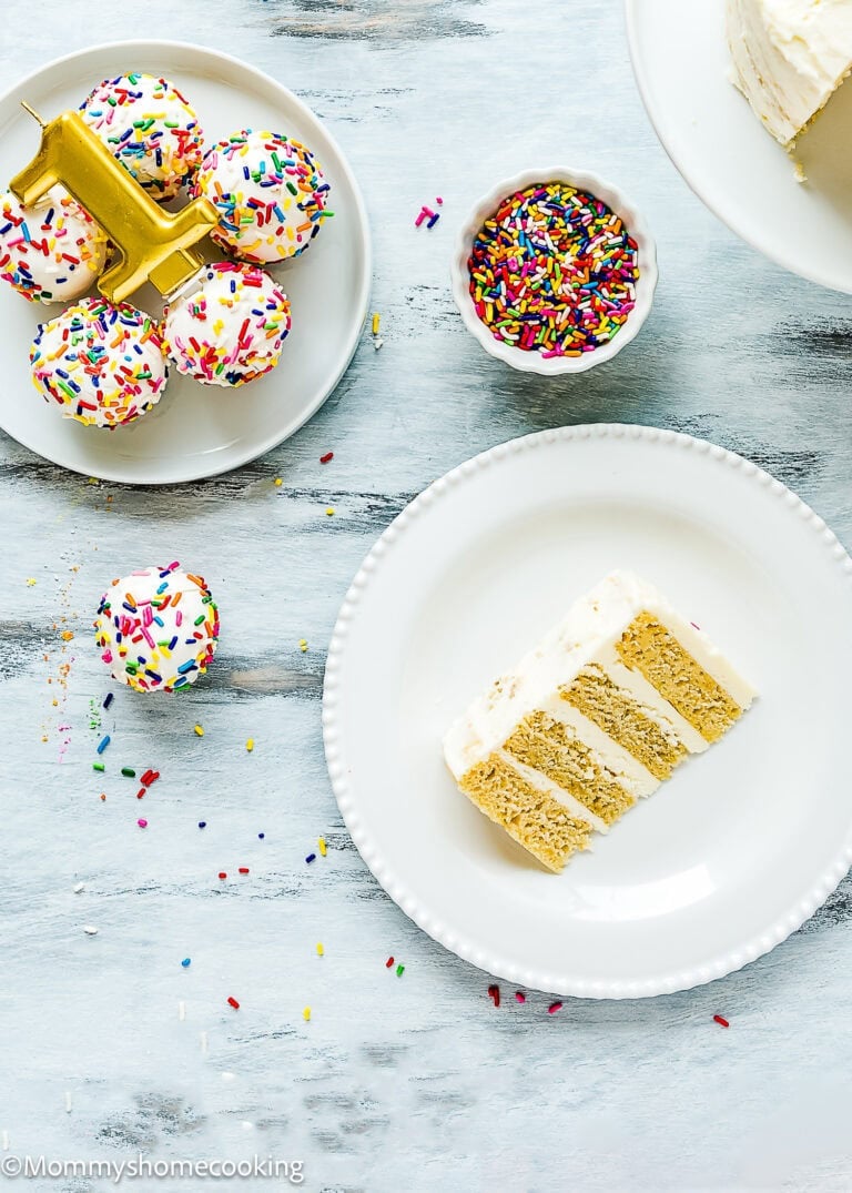 A slice of layered vanilla Easy Eggless Smash Cake with white frosting on a plate, cake pops with sprinkles, and a bowl of rainbow sprinkles on a light surface.
