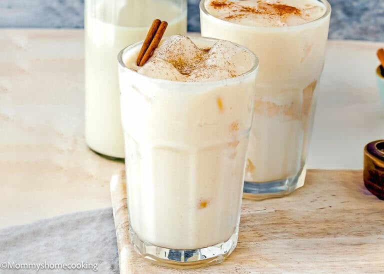 Two glasses of iced Venezuelan Chicha, topped with ground cinnamon and cinnamon sticks, sit on a wooden surface with a bottle of milk in the background.
