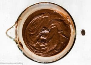 A glass mixing bowl filled with smooth, glossy batter for The Best Eggless Chocolate Cake Ever sits on a white surface.