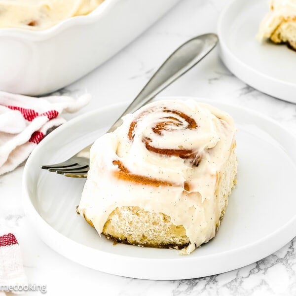 An Easy Eggless Cinnamon Roll with frosting sits on a white plate with a fork beside it; a baking dish and another plate are in the background.