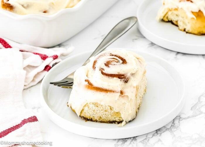 An Easy Eggless Cinnamon Roll with frosting sits on a white plate with a fork beside it; a baking dish and another plate are in the background.