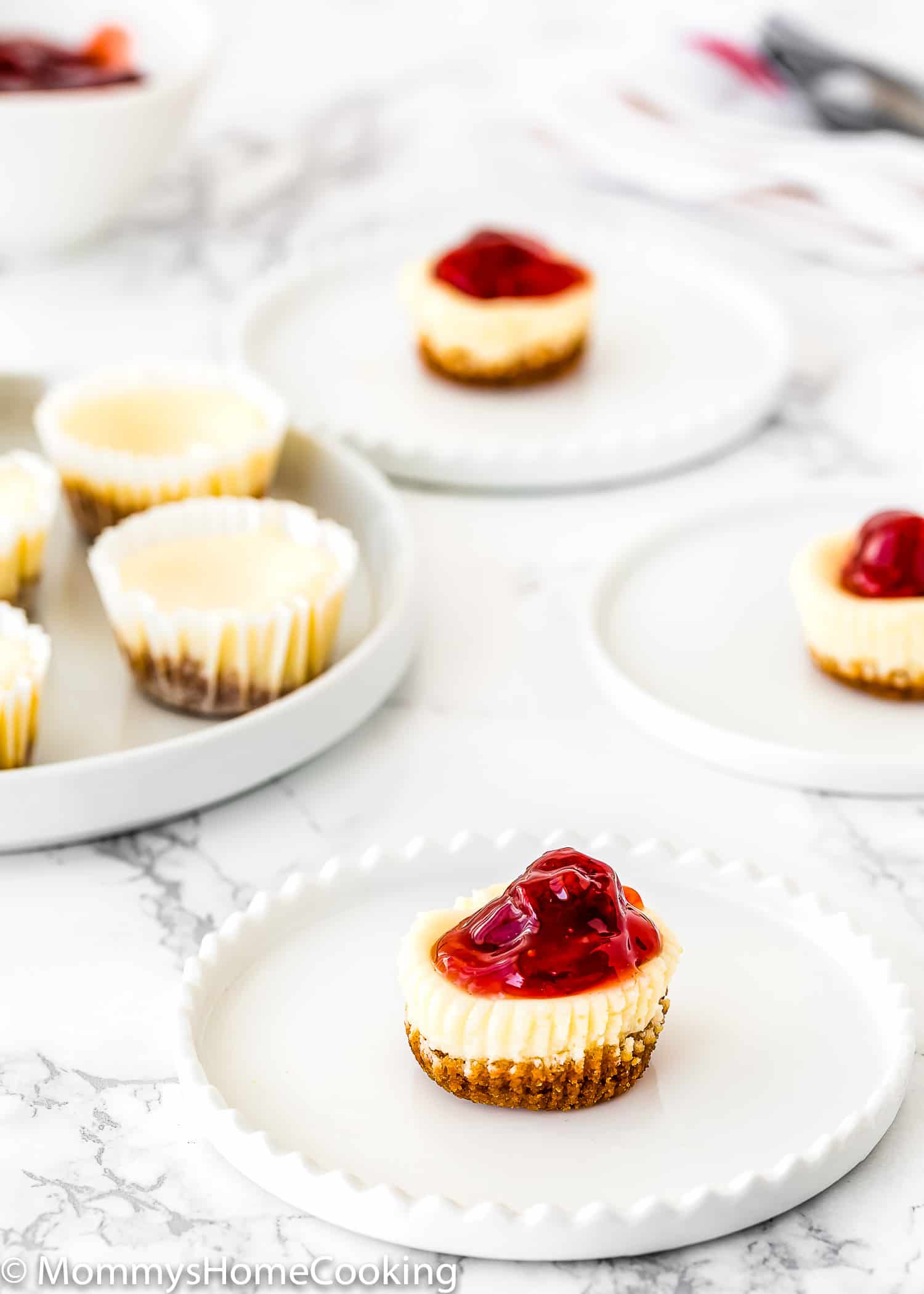 An Eggless Mini Cheesecake with strawberry sauce on top over a white plate with more mini cheesecakes in the background.