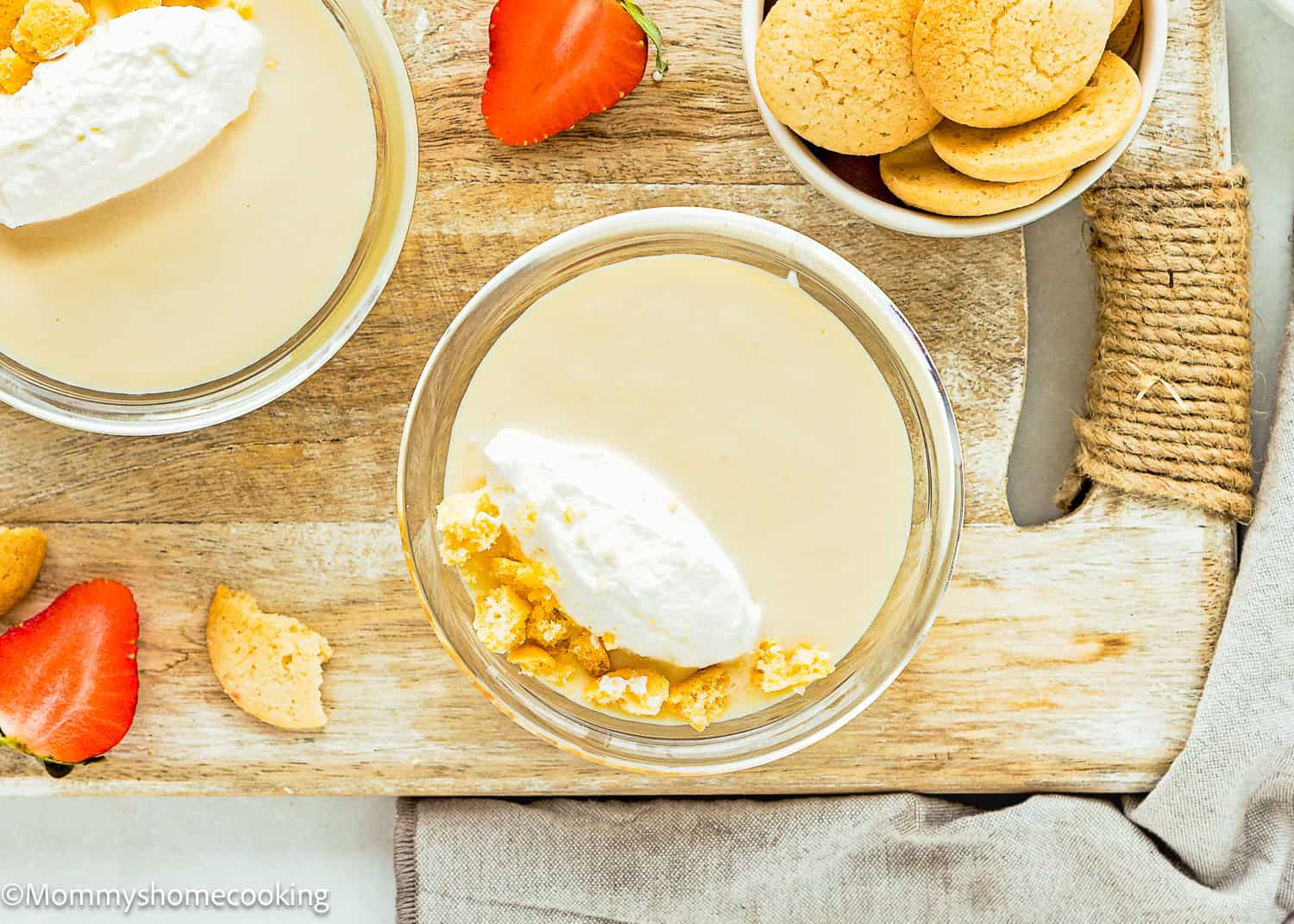 Two bowls of Easy Eggless Vanilla Pudding sit on a wooden board, topped with whipped cream and cookie crumbles. Fresh strawberries and a small bowl of round cookies are arranged alongside for a delightful presentation.