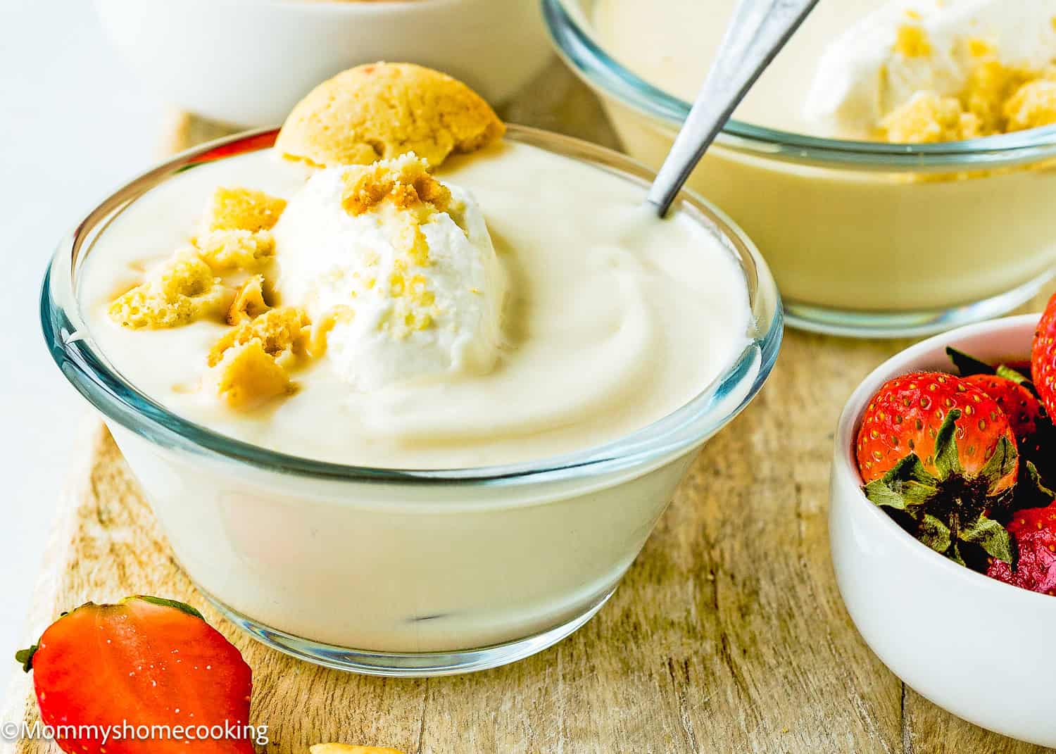 A glass bowl of Easy Eggless Vanilla Pudding topped with whipped cream and crumbled cookies sits on a wooden surface, next to strawberries and another bowl of pudding.