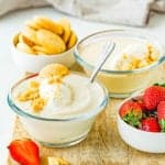 two bowls with Egg free homemade vanilla pudding with whipped cream over a wooden board with fresh strawberries, more vanilla wafers and a gray kitchen towel.