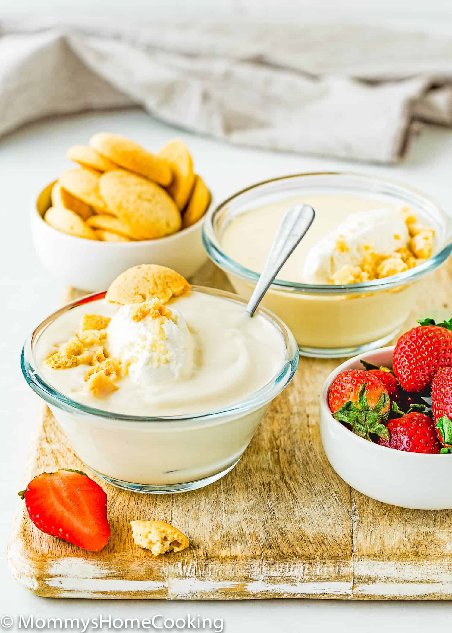 two bowls with Egg free homemade vanilla pudding with whipped cream over a wooden board with fresh strawberries, more vanilla wafers and a gray kitchen towel.
