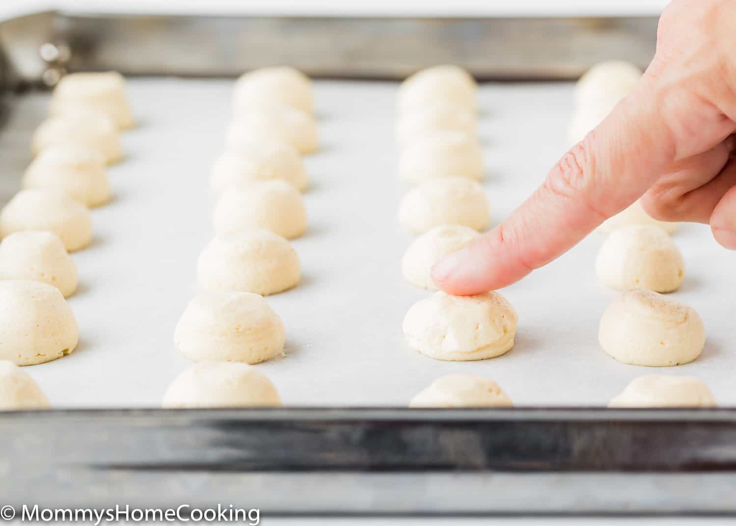 a finger smoothing the tops of Eggless Vanilla Wafers dough scoop out in a baking tray.