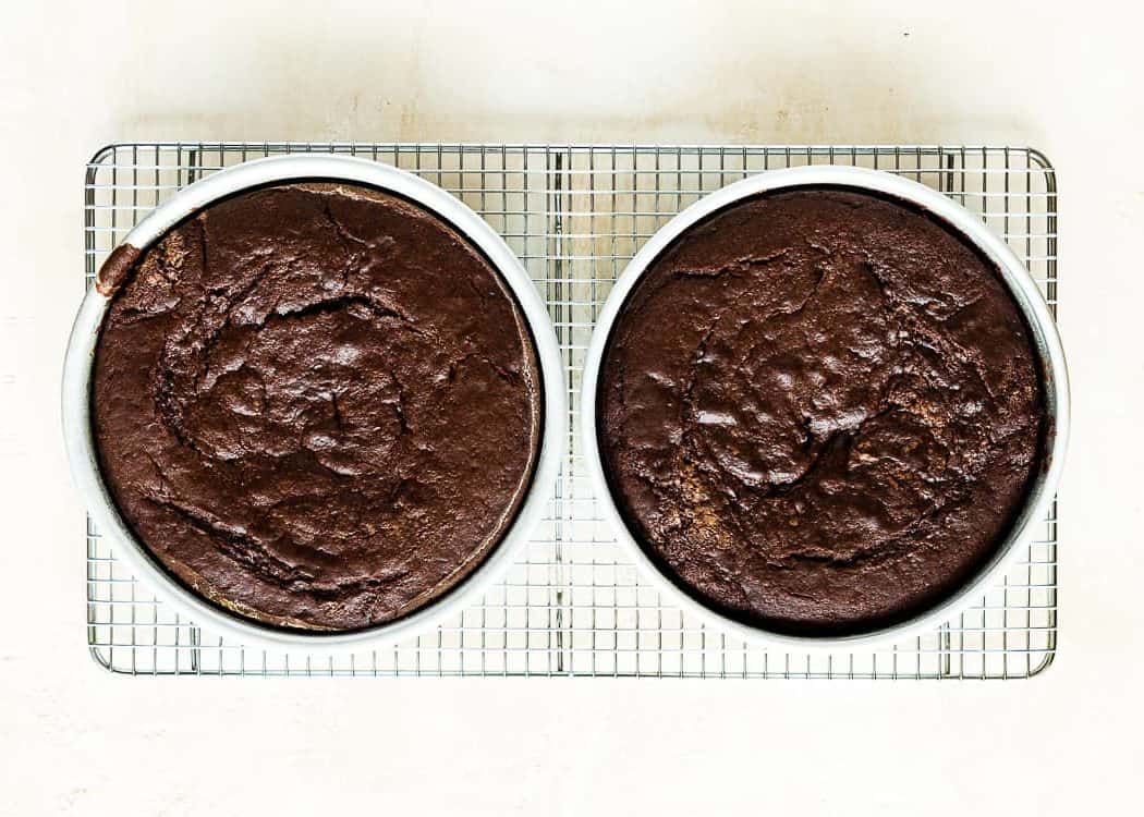 Two cake pans with eggless chocolate cakes over a cooling rack. 