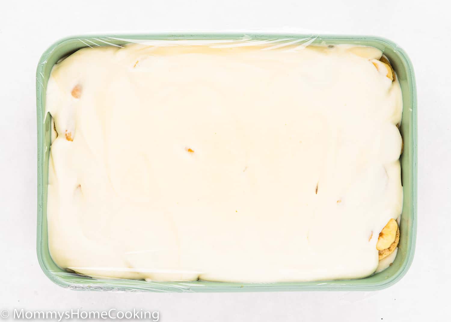 Homemade eggless banana pudding in a green rectangular dish covered with plastic wrap.