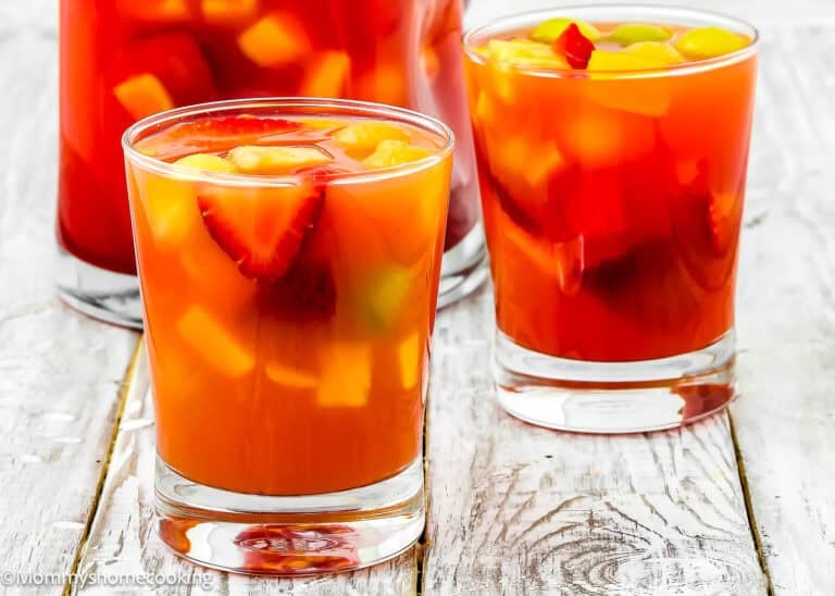 Two glasses of Venezuelan Tizana (Easy Fruit Punch Mocktail) with ice and assorted fruit pieces, including strawberries and peaches, rest on a rustic white wooden surface.