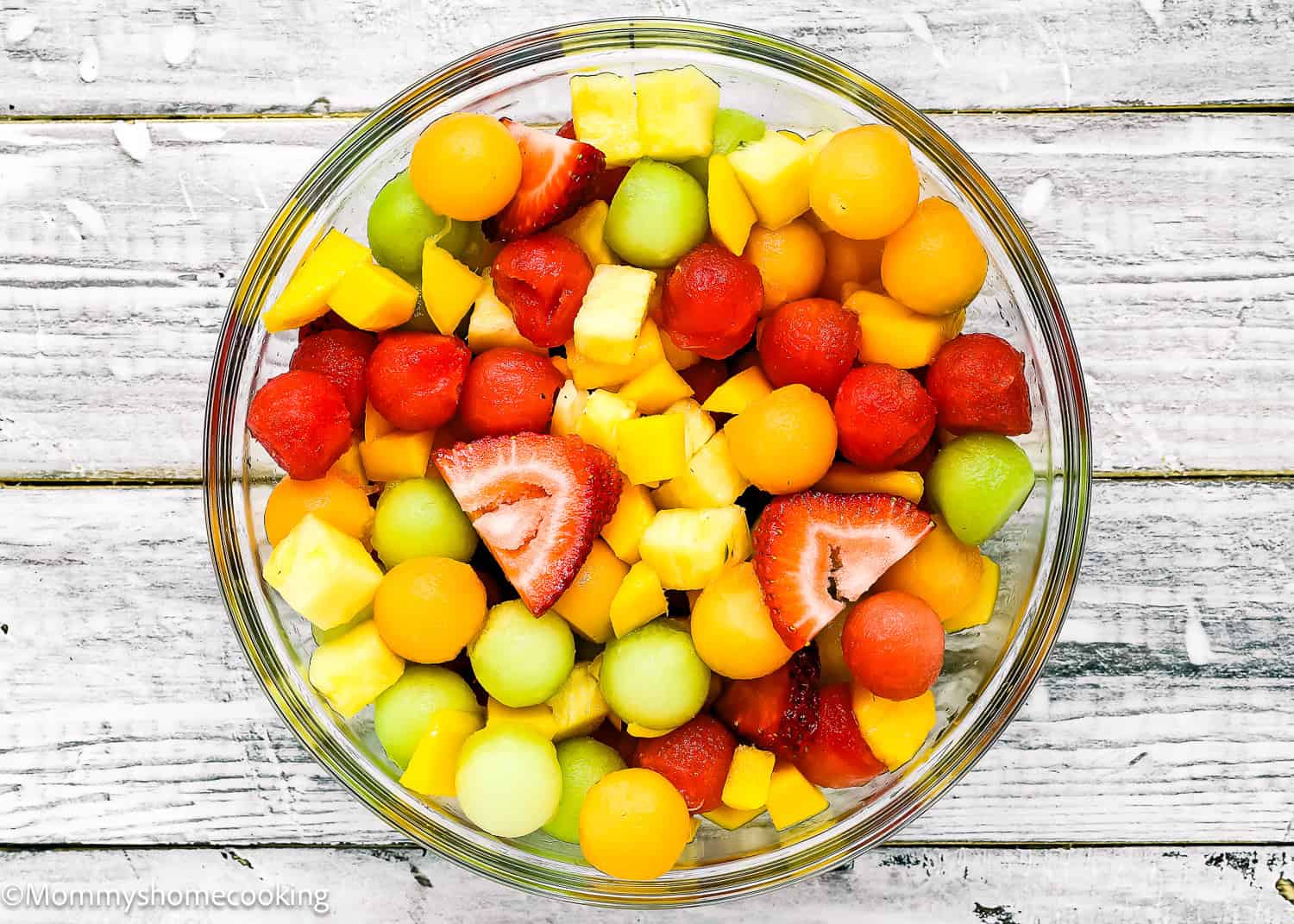 A glass bowl filled with assorted fresh fruit, reminiscent of a Venezuelan Tizana (Easy Fruit Punch Mocktail), including strawberries, melon balls, pineapple chunks, and mango pieces on a white wooden surface.